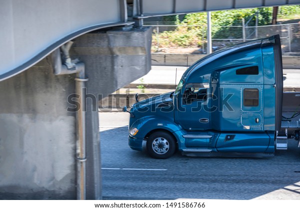 Blue big rig\
American bonnet semi truck with high roof transporting commercial\
cargo on flat bed semi trailer driving on the wide highway road\
under the bridge in sunny\
day