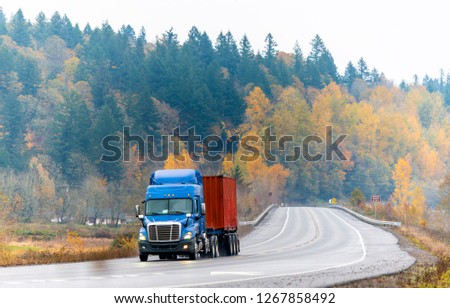 Blue big rig day cab for local haul semi truck with flat bed semi trailer transporting container running on winding autumn road with raining weather with yellow trees on the hill