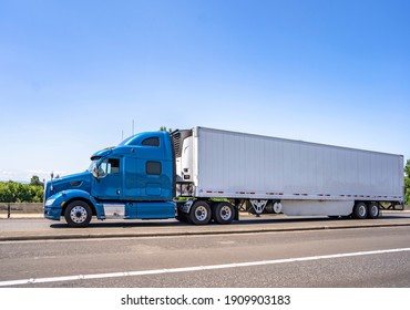 Blue Big Rig Industrial Grade Bonnet Long Hauler Diesel Semi Truck With High Roof Cab And Refrigerator Semi Trailer Running With Commercial Cargo On The Wide Highway Road With Green Trees Hillside