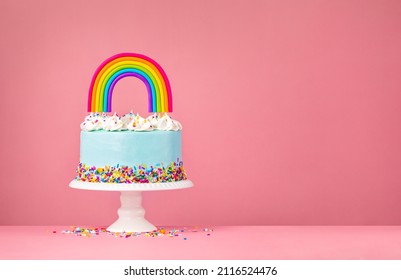 Blue Birthday Cake With Meringue Decorations, Sprinkles And A Colorful Homemade Rainbow Topper Over A Pink Background.