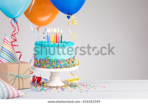 Blue Birthday cake, presents, hats and colorful\
balloons over light grey.