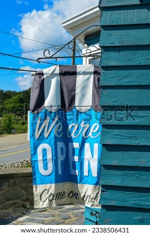 A blue, black, and white colored cloth flag hanging with the text We are open come on in. The banner advertisement is attached outside to the wall of a teal-green wooden cafe shop under blue sky. 