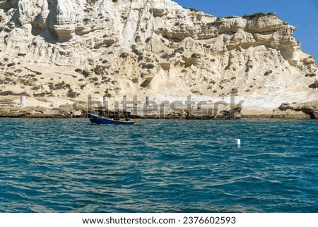 A blue boat is near a rocky cliff, with the deep blue ocean stretching out beyond