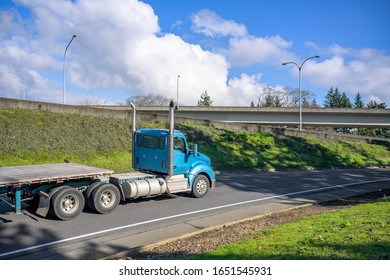 Blue Bonnet Powerful Big Rig Day Cab Semi Truck With Vertical Exhaust Pipes Transporting Empty Flat Bed Semi Trailer Moving On The One Line Road Entrance With Green Hill On The Side