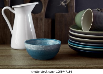 Blue Bowl, Stack Of Plates, Coffee Mugs And White Metal Jar On Rustic Wooden Table Against Shabby Grey Wall With Cutting Boards. Crockery.