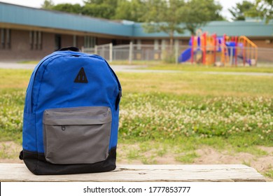 Blue Boy's Backpack On The Old Wooden Bench. Elementary School With Kindergarten Playground In The Background. Back To School Concept. No People