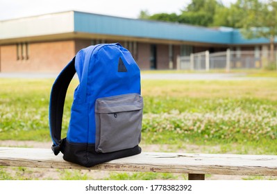 Blue Boy's Backpack On The Old Wooden Bench. Elementary School In The Background. Back To School Concept. No People