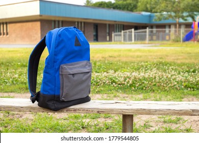 Blue Boy's Backpack On The Old Wooden Bench. Elementary School With Kindergarten Playground In The Background. Back To School Concept. No People