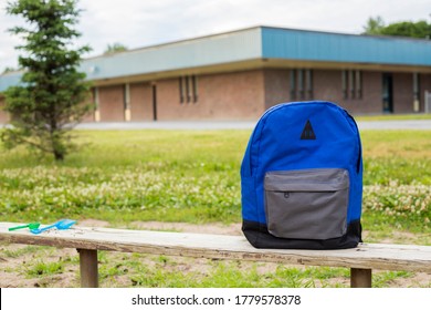 Blue Boy's Backpack On The Old Wooden Bench. Elementary School In The Background. Back To School And Return To School Concept. Small Children's Shovels On The Bench. No People