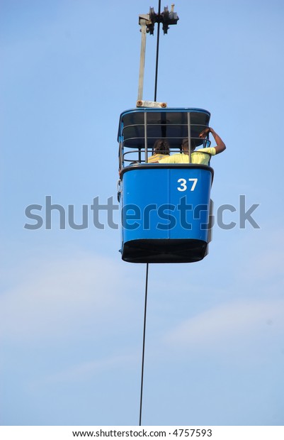 Blue Cable Car from a sky ride high above a theme\
park.  Two people, a man and a woman are visible.  There is a blue\
sky with thin clouds.