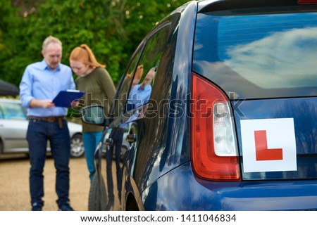 Blue car with a learner driver sign with driving student and instructor in the background