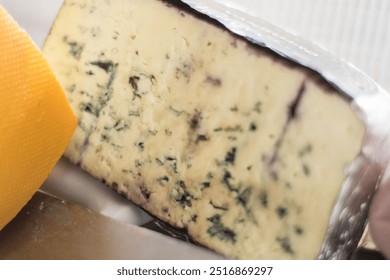 Blue cheese, with a creamy texture and a light color that has streaks of blue and green mold. It is wrapped in aluminum foil. Next to it, a piece of yellow cheese contrasts with the blue cheese. - Powered by Shutterstock