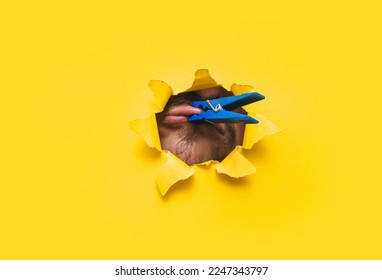 Blue clothespin on the lips of a mute man close up. Torn hole in yellow paper. The concept of a chatty person, dumbness, hate speech and silence. - Shutterstock ID 2247343797