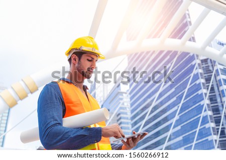 Blue collar worker, engineer man work on digital tablet, hold blue print, wear safety hat, stand near worksite. Worker guy survey a land for big project of building or estate. look determined