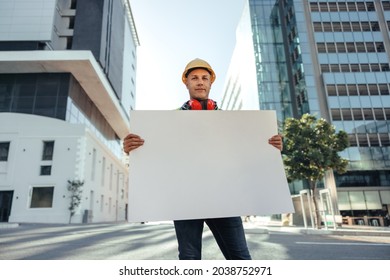 Blue Collar Worker Looking At The Camera While Holding A Blank Placard In The City. Mid-adult Construction Worker Standing In Front Of High Rise Buildings In His Workwear.