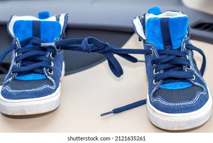 Blue Color Shoes For Kids With Tied Laces, In A Modern Car. Shoes On The Steering Wheel, On The Car's Seat Or In A Boy's Hands. April Fools Day Prank.