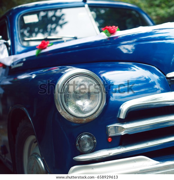 Blue colored vintage wedding car decorated with\
roses and white ribbon. Mag wheels. Hipster style. Daylight. Close\
up. Outdoor shot