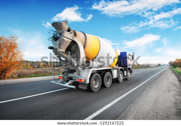 Blue concrete mixer truck on the\
countryside road with trees against blue sky with\
clouds