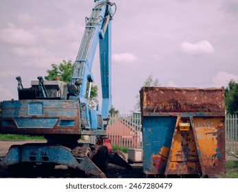 A blue construction machine is parked next to a yellow dumpster. The scene is somewhat bleak and industrial, with the blue and yellow colors contrasting against the gray sky