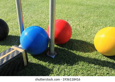 A blue croquet ball is about to be struck with a croquet mallet through a croquet hoop to hit the red ball on a green croquet lawn with the yellow and black balls on either side