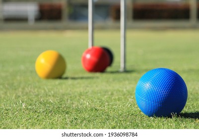 A blue croquet ball is in focus in the foreground of a croquet lawn, while the red, yellow and black balls and the hoop are out of focus in the background