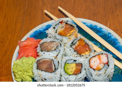 A Blue Decorative Pottery Plate With Six Sushi Rolls And Two Chopsticks Sit On A Wooden Table. The Sushi Includes Tuna, Avocado, Seaweed, Salmon And Rice. There Are Wasabi And A Palette Cleanser.