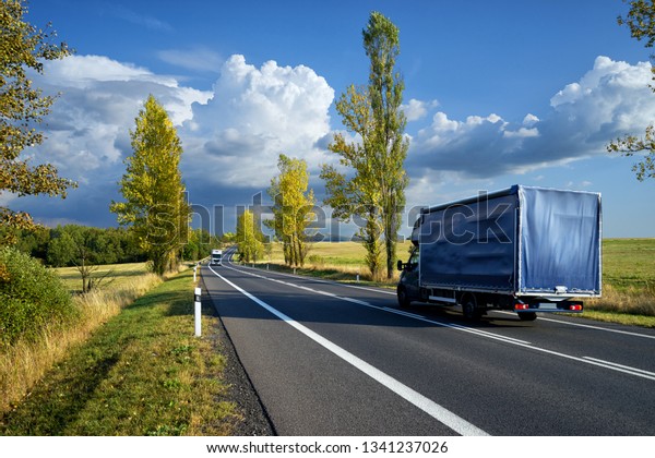 Blue delivery van and white truck driving on the asphalt\
road between line of trees in autumn landscape                     \
         