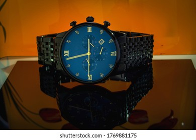 A Blue Dial Watch On A Glassy Surface With Orange Background.