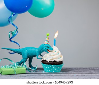 Blue Dinosaur Toy Eating A Birthday Cupcake  On A Gray Background.