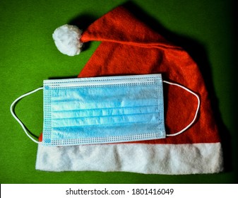 A Blue Disposable Medical Face Mask On A Red Santa Hat On Green Flannel Background In Dim Light. Accessory For Christmas Time Celebration In 2020-2021s. Prevent From COVID-19 During Party In Holiday.