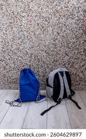 A blue drawstring bag and a gray backpack placed on a wooden floor against a textured pebble wall indoors
