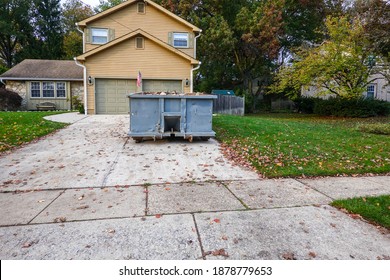 A blue dumpster filled with construction debris in the driveway of a yellow house in front of the garage doors
