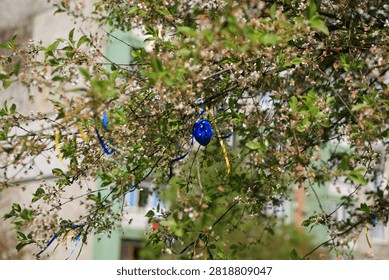 Blue Easter egg hanging on a tree branch, surrounded by small white blossoms and green leaves. Blue and yellow ribbons are also tied to the branches, creating a festive appearance.