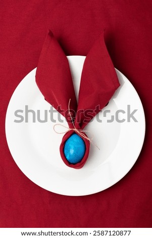 A blue Easter egg and a red napkin as bunny or rabbit ears. Easter celebration concept. Top view vertical photo of colorful easter egg on red textile background