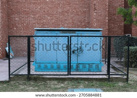 Blue Electrical Transformer Box Protected by Fence against Brick Wall