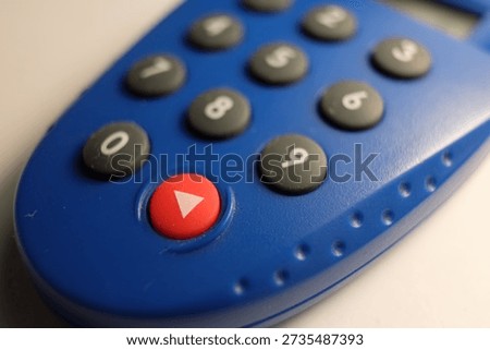 A blue electronic security token featuring a numeric keypad with black circular buttons and a prominent red "play" button. The plastic device has a textured grip and a shallow depth of field.