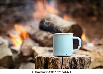 Blue enamel cup of hot steaming coffee sitting on an old log by an outdoor campfire. Selective focus on mug with blurred background.