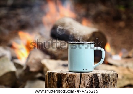 Blue enamel cup of hot steaming coffee sitting on an old log by an outdoor campfire. Selective focus on mug with blurred background.