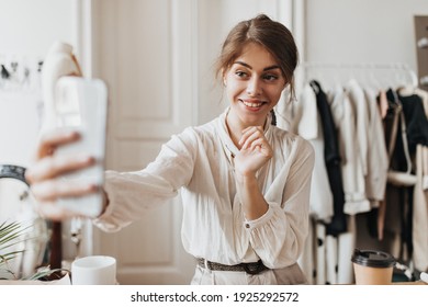 Blue eyed woman in white blouse taking photos. Brunette charming lady in good humore makes selfie, smiles and holds cell-phone - Shutterstock ID 1925292572