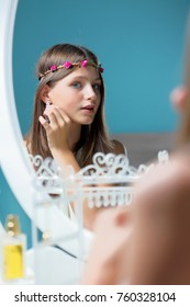 Blue Eyes And Brown Hair Young Smiling Little Girl Kid Teenager With A Crown Of Flowers On Her Head In Front Of A Mirror Learns To Make Up, And Plays With Her Earring