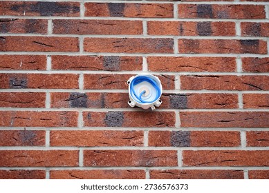 Blue flexible cable and plastic junction box attached to red brick wall. Unfinished security system setup on brick wall, blue network cable coming out of circular white mounting plate. Selective focus