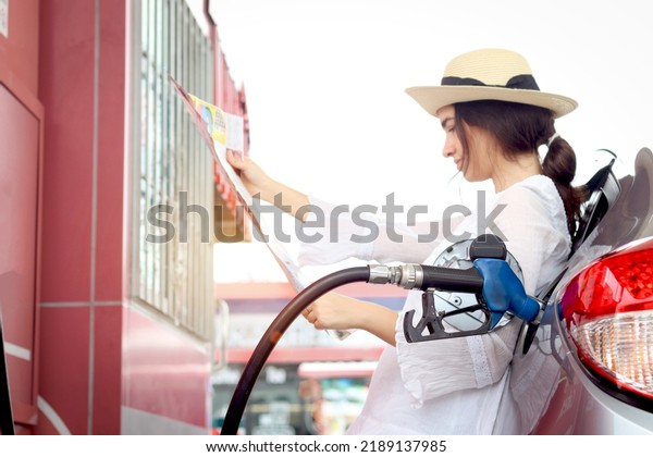 Blue fuel petrol pump nozzle against refuelling car\
with petrol with beautiful female traveler holding touristic map,\
waiting as blurred background at gas station, customer woman\
self-refuelling car