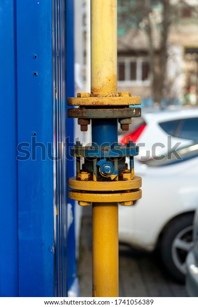 Blue gas crane with flanges on the pipe near\
dark blue metal wall. Gas supply system is distributed gas and turn\
it off if necessary, located in the yard. Pipe painted yellow.\
Vertical orientation.