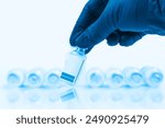 Blue gloved doctor hand holding a vaccine vial in front of a blurred row of vaccine vials on a blue background.