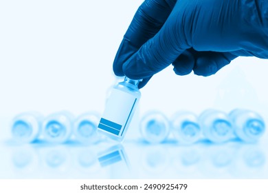 Blue gloved doctor hand holding a vaccine vial in front of a blurred row of vaccine vials on a blue background. - Powered by Shutterstock