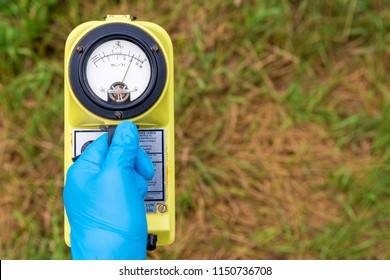 A blue gloved hand holds a radiation meter. The meter shows a high level of radiation. Grass in the background, most of which is brown and dead.