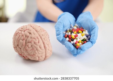 Blue gloved hands present assorted colorful capsules next to detailed brain model on bright table, creating clear medical concept for neuroscience, pharmacology, treatment planning, brain health
