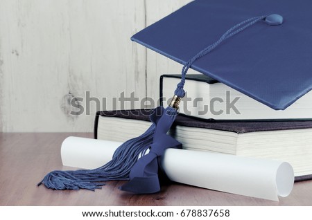 A blue graduation cap resting on two books with a diploma tied with blue ribbon on a wooden background. Copy space. White vignette added