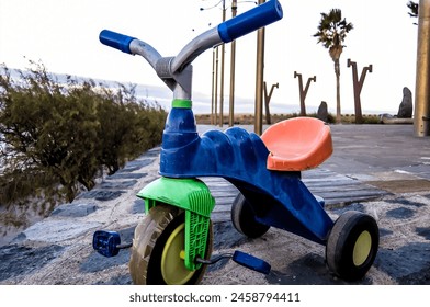 A blue, green and orange tricycle is sitting on a sidewalk near the water. The tricycle is missing a wheel and has a broken handlebar. The scene is peaceful and serene, real image - Powered by Shutterstock