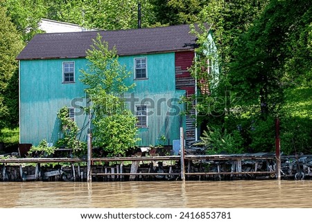 Blue green turquoise house. Shore of river lake water. Trees woods. Disrepair. Windows two story. Sunny day summer. Water's edge. Building home. Metal siding. Dock. 